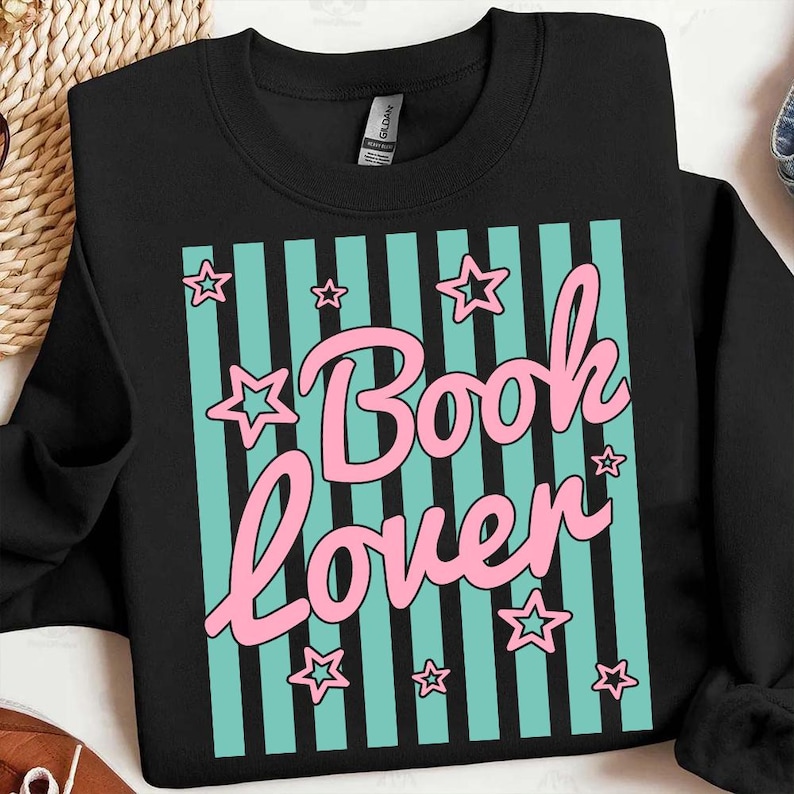 May include: Black sweatshirt with a teal and black striped design. The design features the words "Book Lover" in pink script, surrounded by pink stars. The sweatshirt is made of a soft material.