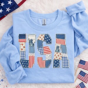 K&ouml;nnte beinhalten: Hellblaues Langarm-Sweatshirt mit dem Wort "USA" auf der Vorderseite. Die Buchstaben bestehen aus Patchwork-Stoff in rot, wei&szlig; und blau, einschlie&szlig;lich Sternen, Streifen und floralen Mustern. Die Freiheitsstatue ist ebenfalls abgebildet.