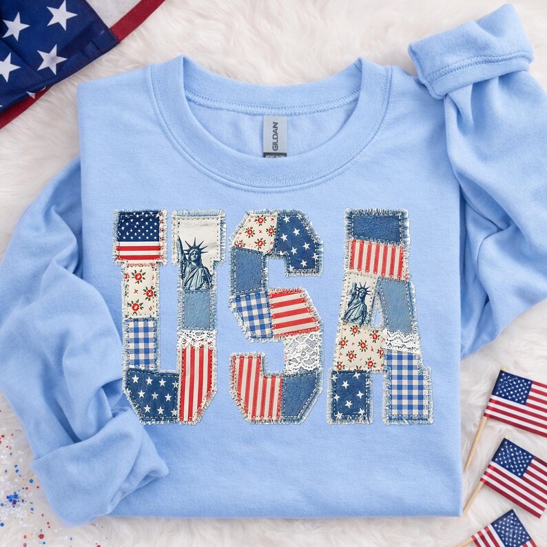 K&ouml;nnte beinhalten: Hellblaues Langarm-Sweatshirt mit dem Wort "USA" auf der Vorderseite. Die Buchstaben bestehen aus Patchwork-Stoff in rot, wei&szlig; und blau, einschlie&szlig;lich Sternen, Streifen und floralen Mustern. Die Freiheitsstatue ist ebenfalls abgebildet.