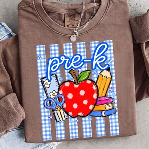 K&ouml;nnte beinhalten: Braunes Sweatshirt mit einem Design f&uuml;r die Vorschule. Das Design umfasst einen roten Apfel, einen Bleistift, B&uuml;cher, eine Schere und die Worte "pre-k" in Blau. Der Hintergrund ist ein blau-wei&szlig;es Vichy-Muster.
