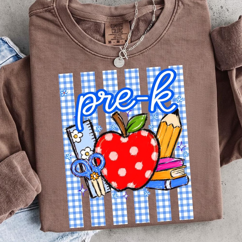 K&ouml;nnte beinhalten: Braunes Sweatshirt mit einem Design f&uuml;r die Vorschule. Das Design umfasst einen roten Apfel, einen Bleistift, B&uuml;cher, eine Schere und die Worte "pre-k" in Blau. Der Hintergrund ist ein blau-wei&szlig;es Vichy-Muster.