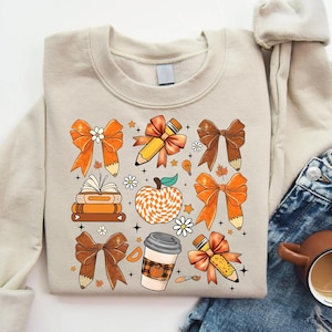 May include: A beige sweatshirt with a pattern of orange and brown bows, pencils, books, a coffee cup, and an apple. The apple is checkered in orange and white.