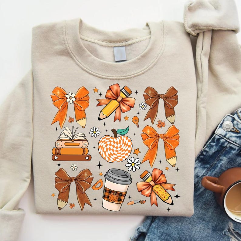 May include: A beige sweatshirt with a pattern of orange and brown bows, pencils, books, a coffee cup, and an apple. The apple is checkered in orange and white.