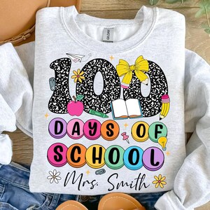May include: A light grey sweatshirt with a colourful graphic design that says "100 Days of School" with a bow, books, pencils, and other school supplies. The text "Mrs. Smith" is at the bottom of the design.