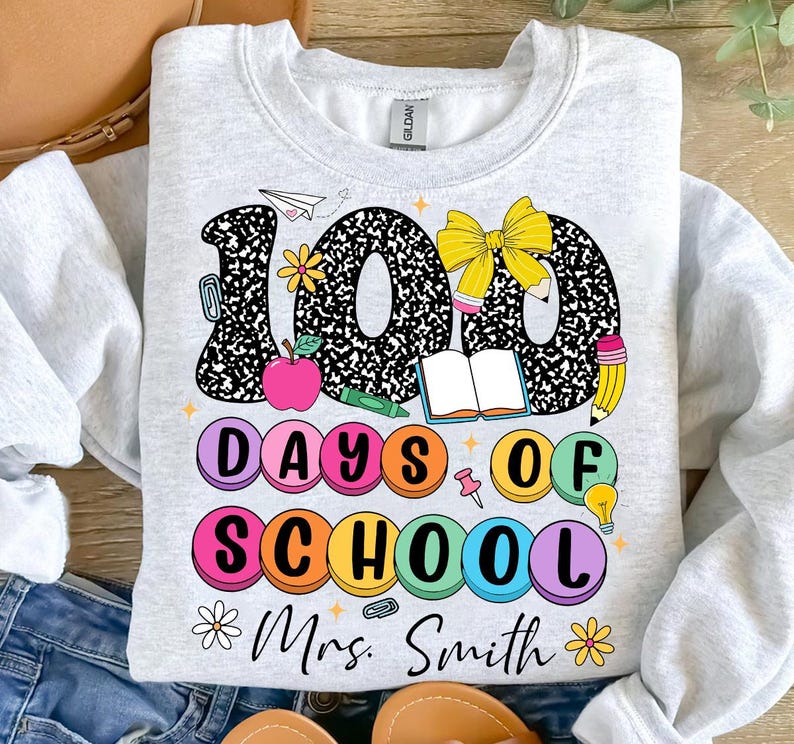 May include: A light grey sweatshirt with a colourful graphic design that says "100 Days of School" with a bow, books, pencils, and other school supplies. The text "Mrs. Smith" is at the bottom of the design.