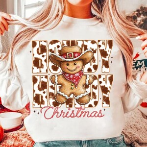 May include: White sweatshirt with a brown and white cow print design. The sweatshirt features a gingerbread man wearing a cowboy hat and the text "Howdy  Christmas".