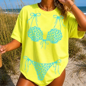 May include: A bright yellow t-shirt with a turquoise bikini design. The bikini top is shaped like seashells with a leopard print pattern, and the bottoms have a matching design. The shirt is a casual, beach-themed garment.