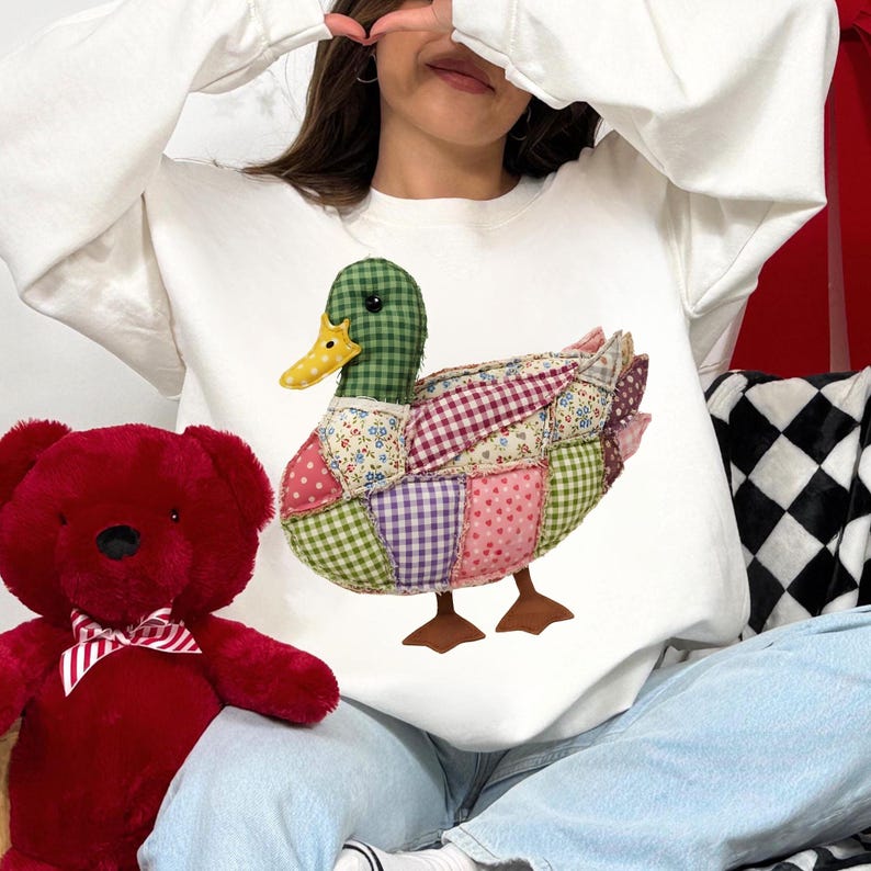 May include: White sweatshirt featuring a patchwork duck design. The duck has a green checkered head, a yellow beak, and a body composed of various fabric patterns including pink polka dots, floral, and gingham. The duck's feet are brown.