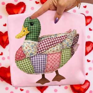 May include: A patchwork duck with a green checkered head and a yellow beak. The duck's body is made of various fabric patterns, including polka dots, floral prints, and gingham. The duck is set against a pink background.