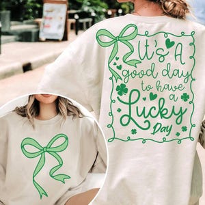 May include: A light beige sweatshirt with a green shamrock design and the text "It's a good day to have a Lucky Day". The sweatshirt also features a green bow on the front.