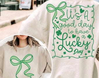 May include: A light beige sweatshirt with a green shamrock design and the text "It's a good day to have a Lucky Day". The sweatshirt also features a green bow on the front.
