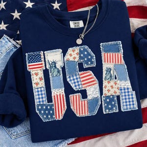 K&ouml;nnte beinhalten: Marineblaues Sweatshirt mit dem Wort "USA" in Patchwork-Design. Das Design zeigt die amerikanische Flagge, die Freiheitsstatue und florale Muster. Eine silberne Halskette mit rundem Anh&auml;nger liegt auf dem Sweatshirt. Marke Comfort Colors.