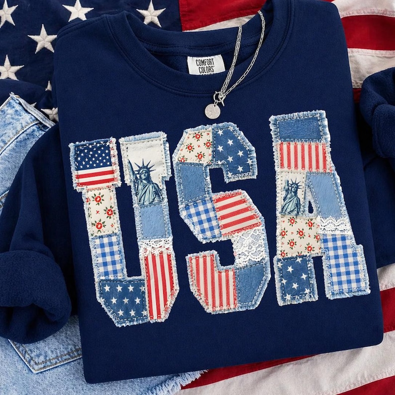 K&ouml;nnte beinhalten: Marineblaues Sweatshirt mit dem Wort "USA" in Patchwork-Design. Das Design zeigt die amerikanische Flagge, die Freiheitsstatue und florale Muster. Eine silberne Halskette mit rundem Anh&auml;nger liegt auf dem Sweatshirt. Marke Comfort Colors.