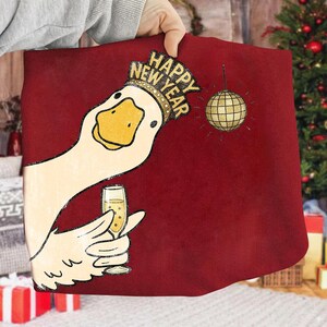 May include: A burgundy pillow with a cartoon goose wearing a gold crown that says "Happy New Year." The goose is holding a champagne glass, and a gold disco ball hangs nearby. The pillow is being held by a person.