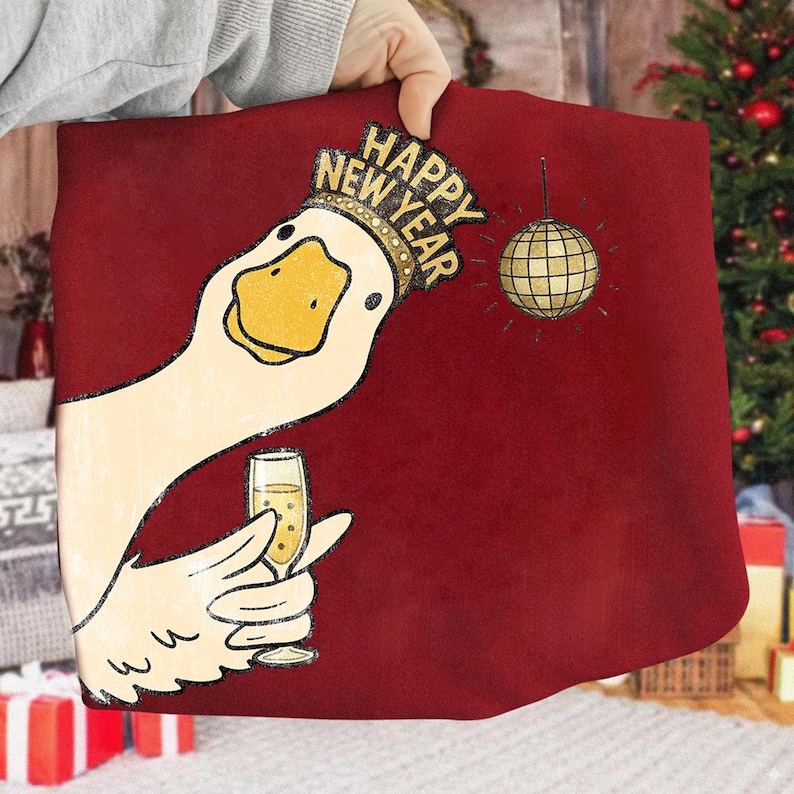 May include: A burgundy pillow with a cartoon goose wearing a gold crown that says "Happy New Year." The goose is holding a champagne glass, and a gold disco ball hangs nearby. The pillow is being held by a person.