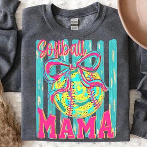 May include: Dark gray sweatshirt with a colorful graphic. The graphic features a softball with a bow, and the words "Softball MAMA" in pink and yellow. The background has vertical teal stripes.