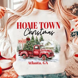 May include: White sweatshirt featuring a red truck carrying a Christmas tree. The text "Home Town Christmas" is printed in red on the sweatshirt. The text "Atlanta, GA" is printed in red below the truck.