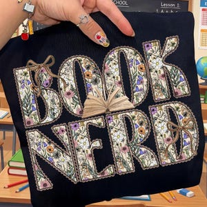 Może przedstawiać: Czarna bluza z napisem "BOOK NERD" w dużych, kwiatowych literach. Litery wypełnione są różnorodnymi kwiatami i obrysowane falistą krawędzią. Mała grafika otwartej książki znajduje się pomiędzy literami. Brązowe kokardy akcentują design.