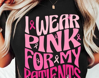 以下が含まれることがあります： ピンクのリボンアクセントが付いたピンクの文字で「I wear pink for my patients」と書かれた黒いTシャツ。