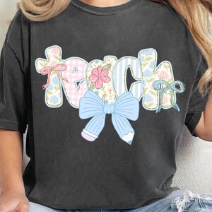 Puede incluir: Una camiseta negra con la palabra "Teach" en letras de patchwork de colores con un lazo azul y detalles de l&aacute;piz.