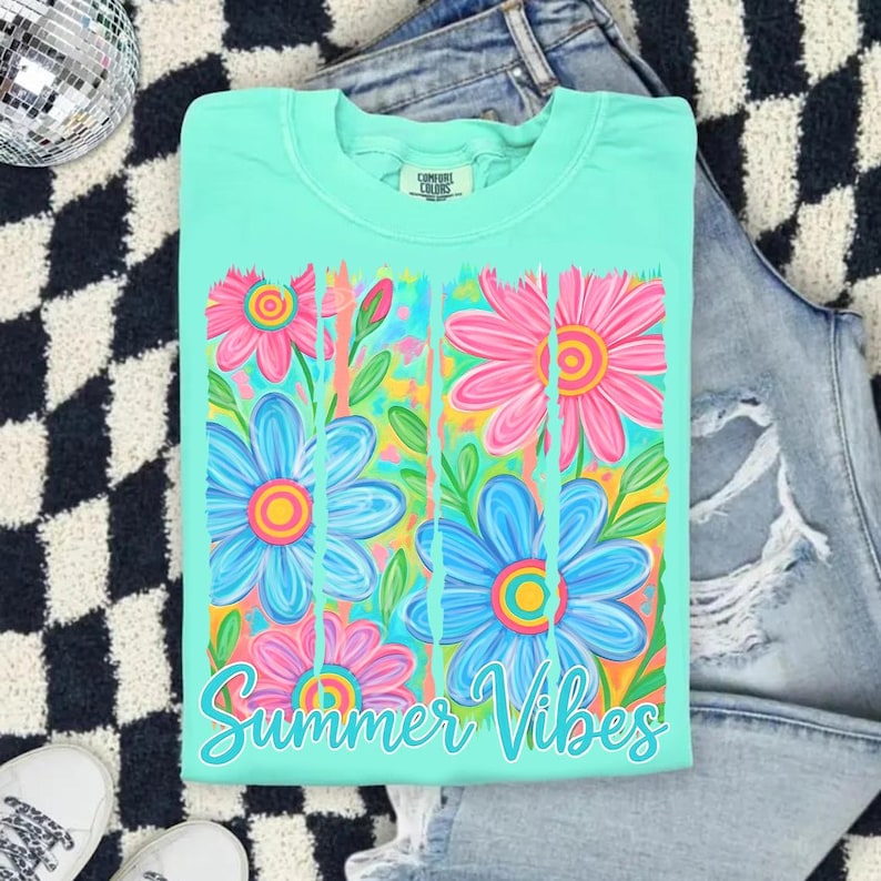 May include: A light teal Comfort Colors t-shirt featuring a floral design with pink and blue flowers and the words "Summer Vibes" in a script font. The shirt is displayed on a black and white checkered surface with a pair of jeans.