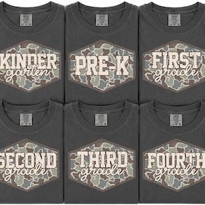 May include: Six charcoal gray t-shirts arranged in two rows. Each shirt features a hexagonal patch with a camouflage pattern and white text reading Kinder garten, Pre-K, First grade, Second grade, Third grade, and Fourth grade respectively.