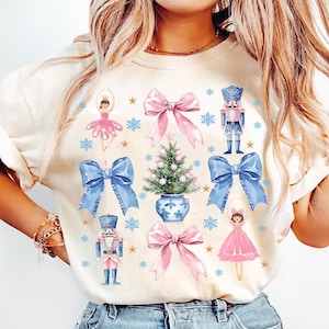 May include: A white t-shirt with a festive Christmas design featuring a ballerina, a nutcracker, a Christmas tree, and bows in pink and blue.