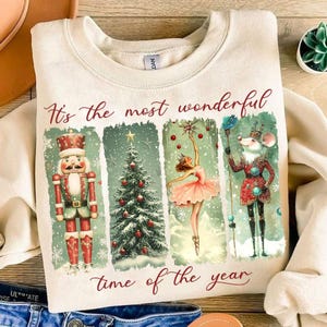 May include: A beige sweatshirt with a Christmas-themed design. The design features a nutcracker, a Christmas tree, a ballerina, and a mouse. The text "It's the most wonderful time of the year" is printed on the sweatshirt.