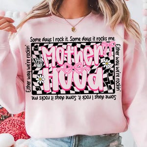 Peut inclure: Sweat-shirt rose clair avec le mot "Motherhood" en grandes lettres roses sur un fond &agrave; carreaux noir et blanc. Le sweat-shirt comporte des n&oelig;uds roses et des marguerites. Le texte "Some days I rock it. Some days it rocks me" est imprim&eacute;.