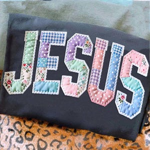 Könnte beinhalten: Dunkelgraues T-Shirt mit dem Wort "JESUS" aus farbenfrohem Patchworkstoff. Die Buchstaben sind mit weißen Nähten umrandet, und der Patchworkstoff enthält Blumen-, Tupfen- und Vichy-Muster.