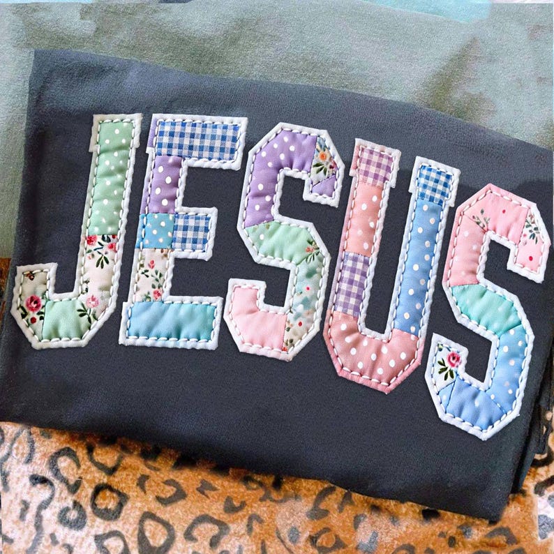 May include: Dark gray t-shirt with the word "JESUS" appliqu&eacute;d in colorful patchwork fabric. The letters are outlined with white stitching, and the patchwork includes floral, polka dot, and gingham patterns.