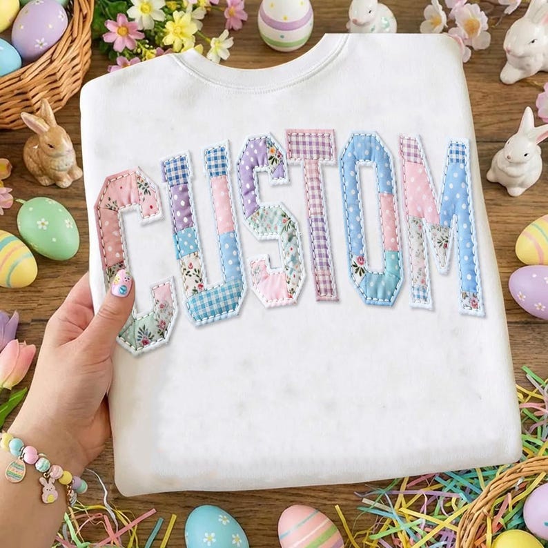 May include: White sweatshirt with the word "CUSTOM" appliqued in patchwork fabric. The letters are in various pastel colors and patterns. The sweatshirt is surrounded by Easter decorations, including eggs and bunnies.