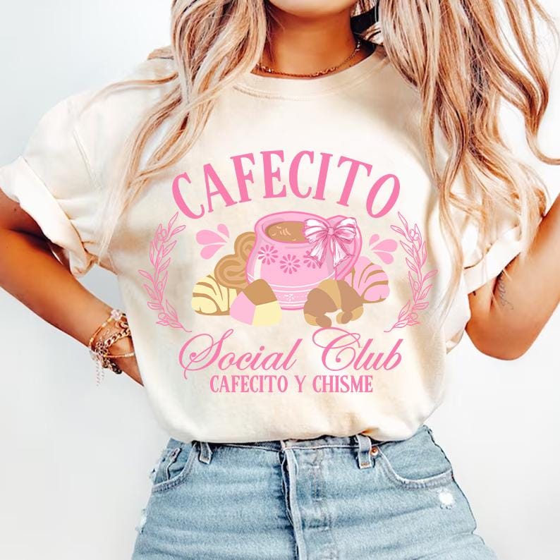 May include: A white t-shirt with a pink graphic design that says "Cafecito Social Club Cafecito y Chisme". The graphic features a pink coffee cup with a bow, surrounded by pastries.