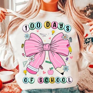 May include: White sweatshirt with a large pink bow graphic and the text "100 Days of School". The design includes school-themed elements like pencils, flowers, and paper airplanes. The text is in a colorful, playful font.