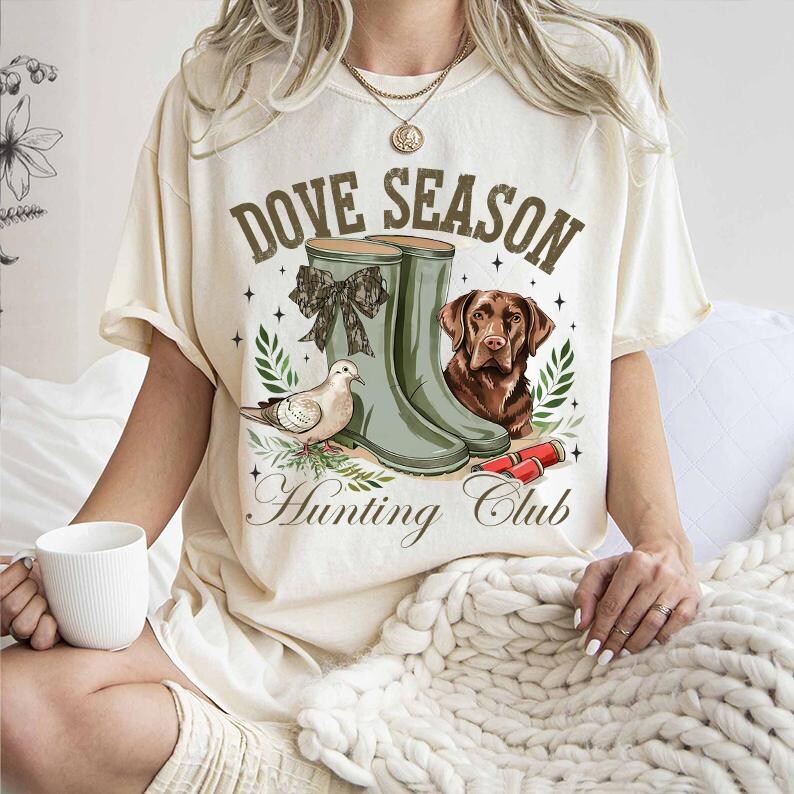 May include: A cream-colored t-shirt with the text "Dove Season Hunting Club" in brown lettering. The shirt features a graphic of two green rubber boots, a dove, and a brown dog.