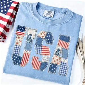 K&ouml;nnte beinhalten: Hellblaues Comfort Colors T-Shirt mit dem aufgen&auml;hten Wort "USA". Die Buchstaben bestehen aus verschiedenen Stoffflicken, darunter die amerikanische Flagge, Blumenmuster und die Freiheitsstatue.