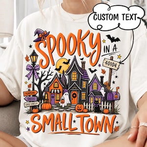 May include: An off-white t-shirt featuring a colourful Halloween graphic with the text SPOOKY IN A SMALL TOWN. The design includes a haunted village, ghosts, pumpkins, a black cat, a street lamp, and a signpost with the text 40104 and CUSTOM TEXT.