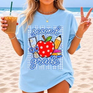 K&ouml;nnte beinhalten: Hellblaues T-Shirt mit einem Grafikdesign. Das Design zeigt einen roten Apfel mit wei&szlig;en Punkten, einen gelben Bleistift und die Worte "second grade" in blauer Schrift. Das Design enth&auml;lt auch Schulmaterial und ein blau-wei&szlig;es Karomuster.
