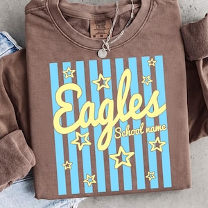 Puede incluir: Una sudadera marr&oacute;n con la palabra "Eagles" en escritura amarilla, sobre un fondo a rayas azul claro con acentos de estrellas amarillas. Las palabras "School name" est&aacute;n debajo de la palabra "Eagles". Se ve un collar plateado.