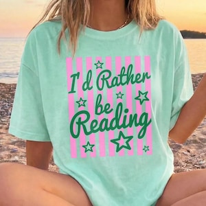 May include: A light green t-shirt with the text "I'd Rather be Reading" in green script and block letters, with pink vertical stripes and green star accents. The shirt is a casual, oversized style.