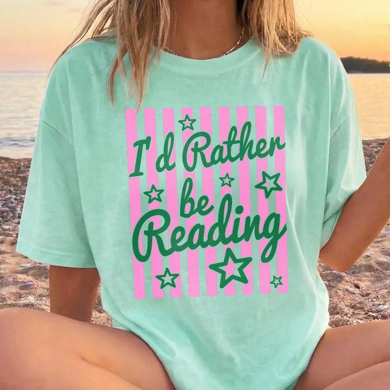May include: A light green t-shirt with the text "I'd Rather be Reading" in green script and block letters, with pink vertical stripes and green star accents. The shirt is a casual, oversized style.