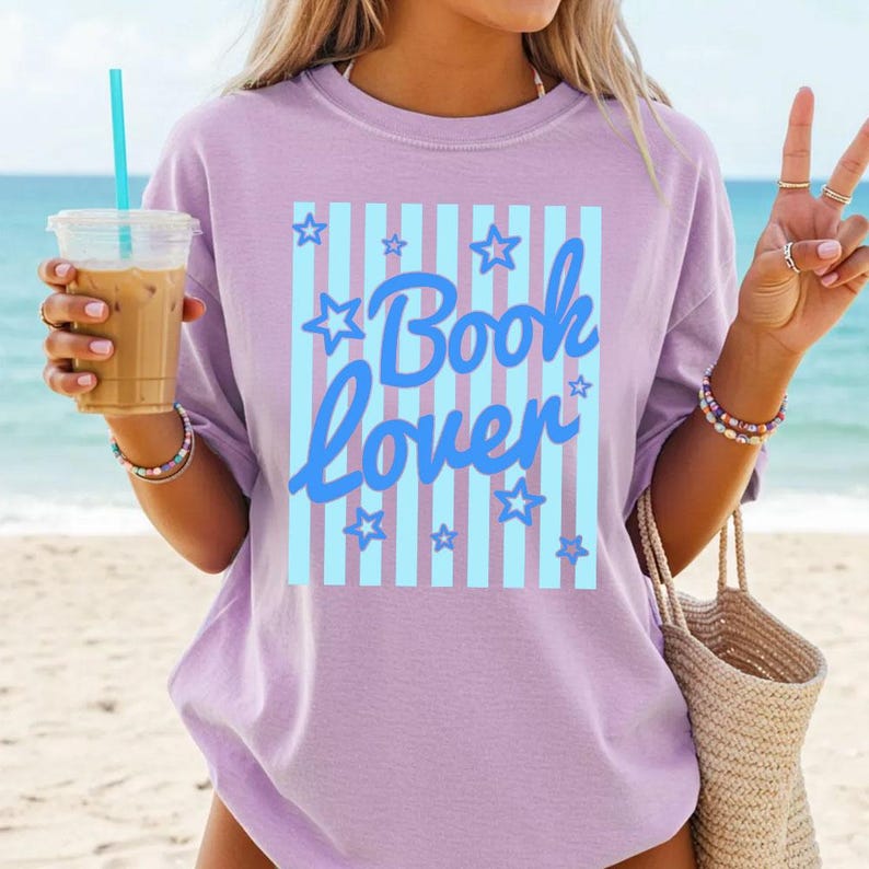 May include: A lilac-colored t-shirt with the words "Book Lover" in blue script, set against a light blue striped background with stars. The shirt is worn by a person holding a drink and a woven tote bag.