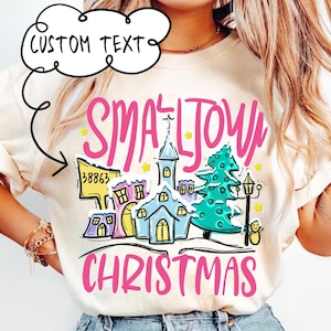 Puede incluir: Una camiseta blanca con una ilustración rosa y azul de un pueblo pequeño con una iglesia, un árbol de Navidad y un muñeco de nieve. El texto "Small Town Christmas" está escrito en letras rosas.
