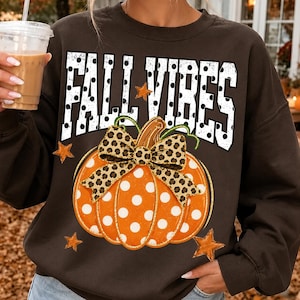 May include: A dark brown crewneck sweatshirt featuring the text FALL VIBES in white with black polka dots. Below the text is an orange pumpkin with white polka dots, a leopard print bow, and several small orange stars scattered around the design.