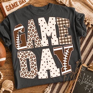 May include: A charcoal gray short-sleeve t-shirt featuring the text GAME DAY in large block letters. The letters are decorated with brown and white patterns including leopard print, polka dots, brown and white plaid, and football graphics.
