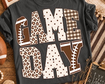 May include: A charcoal gray short-sleeve t-shirt featuring the text GAME DAY in large block letters. The letters are decorated with brown and white patterns including leopard print, polka dots, brown and white plaid, and football graphics.