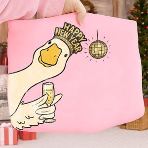 May include: Pink pillow with a cartoon goose wearing a "Happy New Year" crown and holding a champagne glass. A gold disco ball hangs from above. The goose has a yellow beak and is set against a pink background.