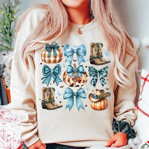 May include: A cream-colored sweatshirt with a fall-themed design featuring pumpkins, bows, and cowboy boots. The pumpkins are decorated with plaid, leopard print, and rhinestones. The bows are blue with white polka dots. The cowboy boots are brown with blue embellishments.