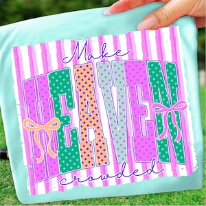 Puede incluir: Camiseta azul claro con un gráfico colorido que dice "Make HEAVEN crowded". El texto está en letras grandes con lunares rosas, verdes y naranjas, sobre un fondo a rayas. El diseño incluye lazos.