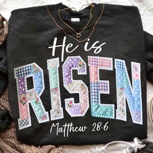 Puede incluir: Sudadera negra con las palabras "He is RISEN" en un dise&ntilde;o de patchwork. Las letras est&aacute;n hechas de varias telas pastel. El texto "Matthew 28:6" est&aacute; debajo de la palabra "RISEN". Un collar dorado est&aacute; drapeado sobre la sudadera.